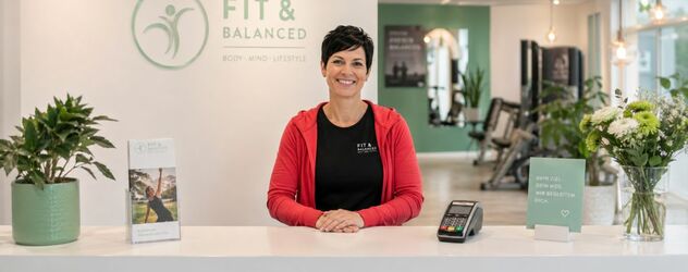 eine Faszientrainerin steht lächelnd hinter dem Empfangstresen eines Fitnessstudios samt EC-Kartenlesegerät