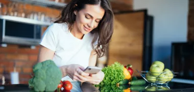 eine junge Frau stützt sich an der Küchenzeile, auf dem verschiedene Gemüsesorten liegen und schaut auf ihren Handy