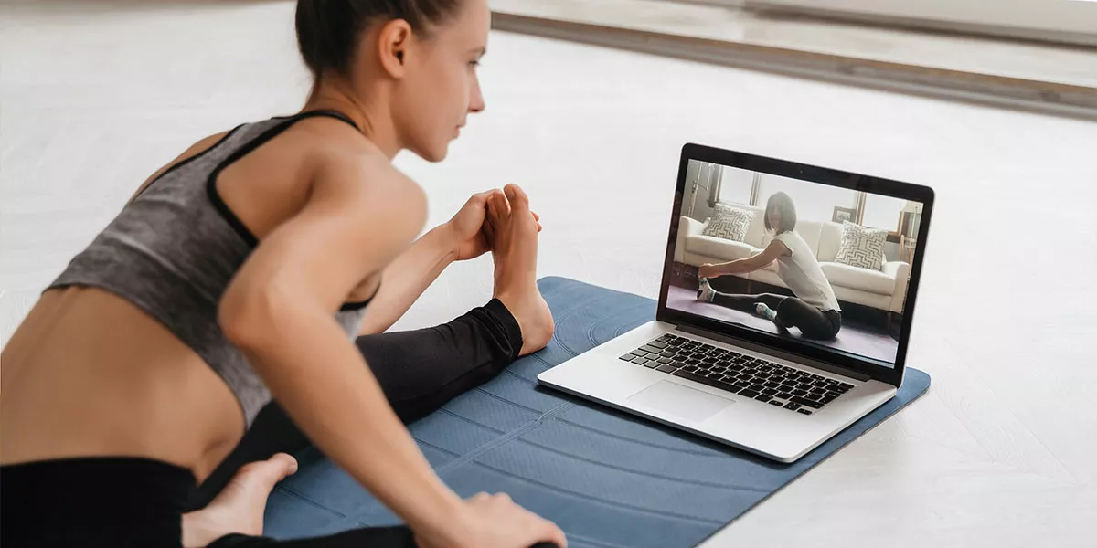 Eine Frau führt vor dem Laptop auf einer Matte sitzend Pilatesübungen aus